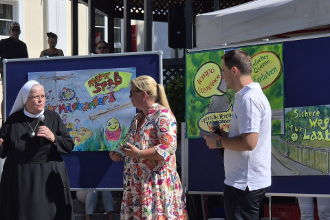 Ordensfrau spricht zu Besuchern, daneben eine Frau und ein Mann vor Plakaten.