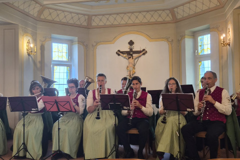 Musikerinnen und Musiker in Tracht spielen in einem Raum mit Kruzifix vor der Wand.