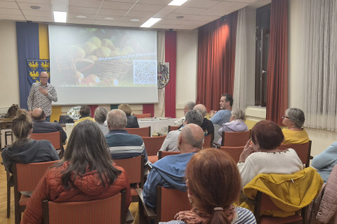 Vortragender steht vor Leinwand mit Pilzkörben, Publikum sitzt im Saal